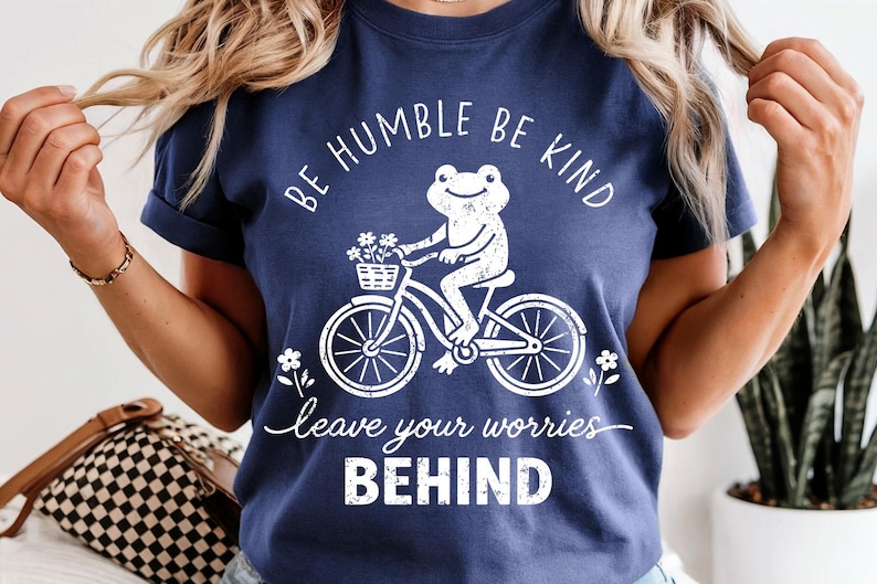 May include: Navy blue t-shirt featuring a white frog on a bicycle, with a basket of flowers. The text says "Be Humble Be Kind" and "Leave Your Worries Behind". A relaxed fit, casual tee.