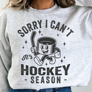 May include: Pale grey sweatshirt featuring a black and white cartoon hockey puck holding a hockey stick. The text says "SORRY I CAN'T It's HOCKEY SEASON". The design includes star shapes.