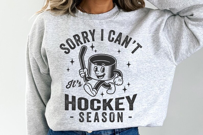 May include: Pale grey sweatshirt featuring a black and white cartoon hockey puck holding a hockey stick. The text says "SORRY I CAN'T It's HOCKEY SEASON". The design includes star shapes.