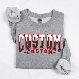 May include: A gray sweatshirt with the word "CUSTOM" in a bold, distressed font. The text is white with a black and red outline. The sleeves are cuffed, and the sweatshirt is laid flat on a white surface.