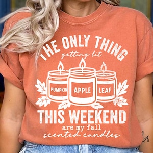 May include: Orange t-shirt with the text "THE ONLY THING getting lit THIS WEEKEND are my fall scented candles." The graphic features three lit candles labeled "Pumpkin," "Apple," and "Leaf."