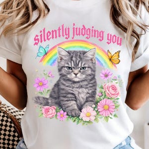 Pu&ograve; includere: T-shirt bianco con illustrazione di un gatto tabby grigio e la scritta "silently judging you". Il design include un arcobaleno, farfalle ed elementi floreali. Una maglietta grafica casual.