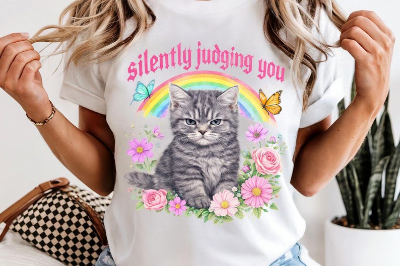 Pu&ograve; includere: T-shirt bianco con illustrazione di un gatto tabby grigio e la scritta "silently judging you". Il design include un arcobaleno, farfalle ed elementi floreali. Una maglietta grafica casual.