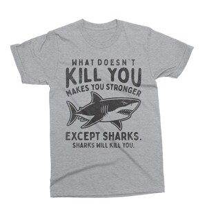 May include: A heather gray t-shirt with the text "WHAT DOESN'T KILL YOU MAKES YOU STRONGER" above a black and white shark illustration. Below the shark, the text reads "EXCEPT SHARKS. SHARKS WILL KILL YOU."