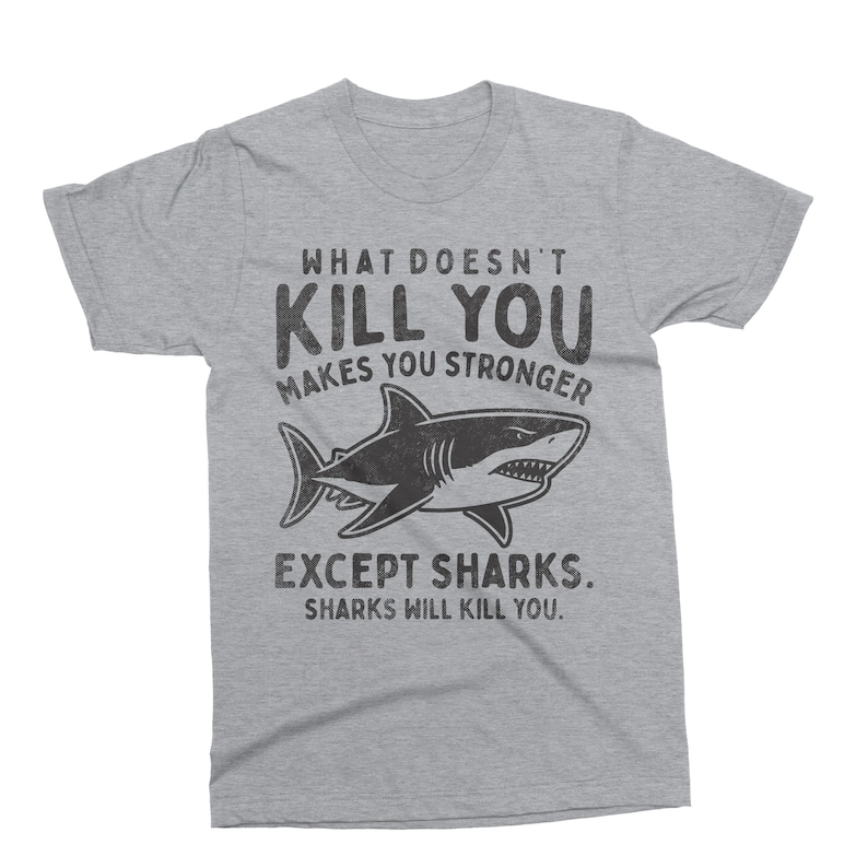 May include: A heather gray t-shirt with the text "WHAT DOESN'T KILL YOU MAKES YOU STRONGER" above a black and white shark illustration. Below the shark, the text reads "EXCEPT SHARKS. SHARKS WILL KILL YOU."