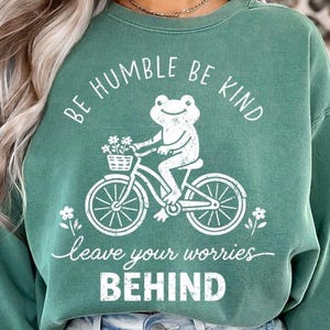 May include: Green sweatshirt featuring a white graphic of a frog on a bicycle with a flower basket. The text reads "BE HUMBLE BE KIND" and "leave your worries BEHIND".