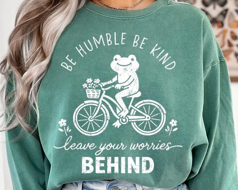 May include: Green sweatshirt featuring a white graphic of a frog on a bicycle with a flower basket. The text reads "BE HUMBLE BE KIND" and "leave your worries BEHIND".