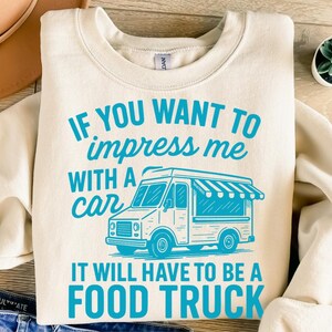 May include: Cream-colored sweatshirt with the text "IF YOU WANT TO impress me WITH A car IT WILL HAVE TO BE A FOOD TRUCK" in turquoise. A turquoise food truck graphic is also featured. The sweatshirt is folded on a wooden surface.