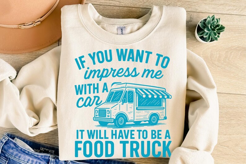 May include: Cream-colored sweatshirt with the text "IF YOU WANT TO impress me WITH A car IT WILL HAVE TO BE A FOOD TRUCK" in turquoise. A turquoise food truck graphic is also featured. The sweatshirt is folded on a wooden surface.