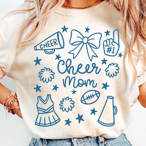 May include: Cream-colored t-shirt with a blue graphic design featuring a megaphone, bow, pom-poms, football, and the words "Cheer Mom." The design includes stars and a cheerleader uniform.