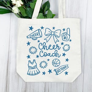 May include: A cream-colored tote bag with a blue design featuring a megaphone, bow, pom-poms, stars, a cheerleading uniform, a basketball, and the words "Cheer Coach" and "Go Team". The bag has white handles and is filled with white flowers and green leaves.