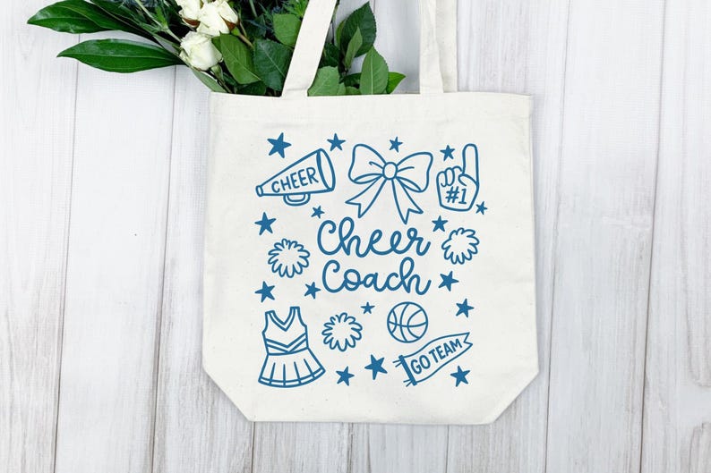 May include: A cream-colored tote bag with a blue design featuring a megaphone, bow, pom-poms, stars, a cheerleading uniform, a basketball, and the words "Cheer Coach" and "Go Team". The bag has white handles and is filled with white flowers and green leaves.