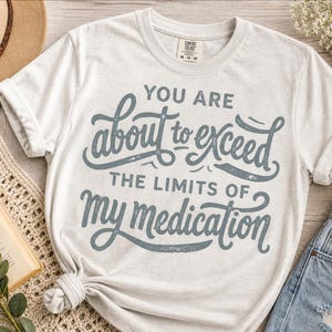 May include: A light gray t-shirt with the text "YOU ARE about to exceed THE LIMITS OF my medication" in a gray script. The shirt is styled with a hat, book, coffee, and jeans.