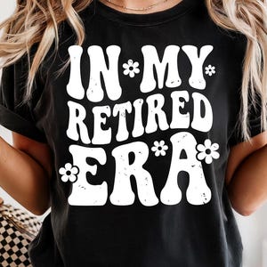 Könnte beinhalten: Schwarzes T-Shirt mit dem weißen Text "IN MY RETIRED ERA" in Retro-Schrift, verziert mit Blumengrafiken. Ein lässiges T-Shirt, geeignet für den Alltag.