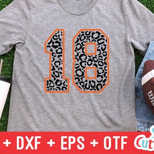 Puede incluir: Una camiseta gris con un gran n&uacute;mero 18 de contorno naranja. Los n&uacute;meros est&aacute;n rellenos con un patr&oacute;n de estampado de leopardo blanco y negro.