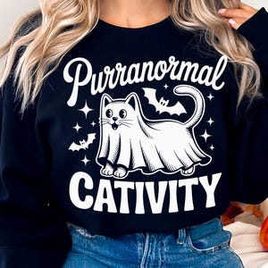 May include: Black sweatshirt with a white graphic of a cat in a ghost costume, bats, and stars. The text reads "Purranormal CATivity" in a playful font, perfect for Halloween.
