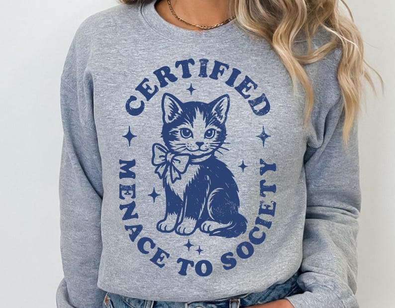 May include: A gray sweatshirt featuring a blue illustration of a cat wearing a bow tie, encircled by the text "CERTIFIED MENACE TO SOCIETY." The design includes stars around the cat and text, creating a vintage aesthetic.