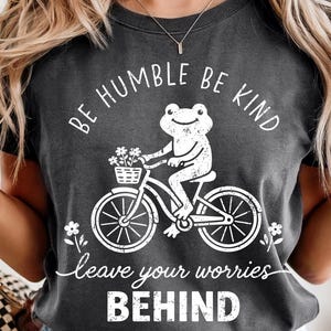 May include: Dark grey t-shirt featuring a white frog on a bicycle, with a basket of flowers. The text says "BE HUMBLE BE KIND" and "leave your worries BEHIND". A casual, comfortable design.