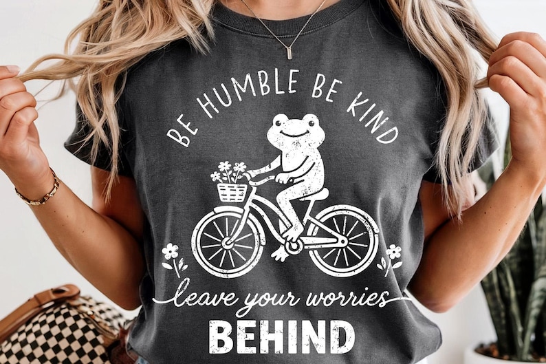 May include: Dark grey t-shirt featuring a white frog on a bicycle, with a basket of flowers. The text says "BE HUMBLE BE KIND" and "leave your worries BEHIND". A casual, comfortable design.