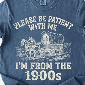 May include: Blue t-shirt with a graphic of a covered wagon pulled by two horses. The text reads "PLEASE BE PATIENT WITH ME" above the wagon and "I'M FROM THE 1900s" below. The shirt has rolled-up sleeves.