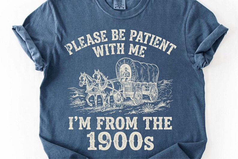 May include: Blue t-shirt with a graphic of a covered wagon pulled by two horses. The text reads "PLEASE BE PATIENT WITH ME" above the wagon and "I'M FROM THE 1900s" below. The shirt has rolled-up sleeves.