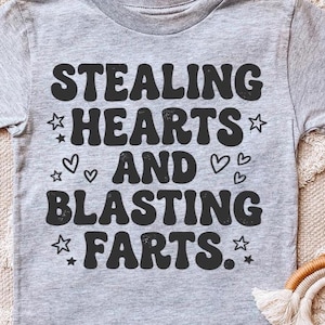 May include: A heather gray t-shirt with the text "STEALING HEARTS AND BLASTING FARTS" in a bold, black font. The text is surrounded by stars and hearts.