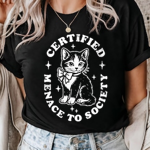 May include: Black t-shirt featuring a black and white cat illustration with a bow tie. The text "CERTIFIED MENACE TO SOCIETY" is printed in a white oval around the cat. The shirt is a crew neck.