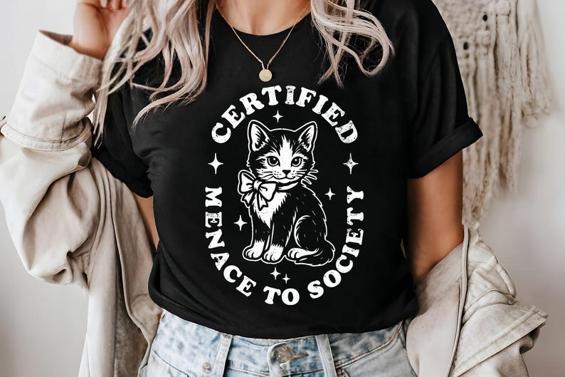 May include: Black t-shirt featuring a black and white cat illustration with a bow tie. The text "CERTIFIED MENACE TO SOCIETY" is printed in a white oval around the cat. The shirt is a crew neck.