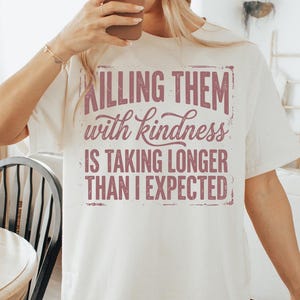 K&ouml;nnte beinhalten: Cremefarbenes T-Shirt mit dem Text "Killing them with kindness is taking longer than I expected" in einer verblassten, malvenfarbenen Schrift. Ein l&auml;ssiges Kleidungsst&uuml;ck f&uuml;r den Alltag.