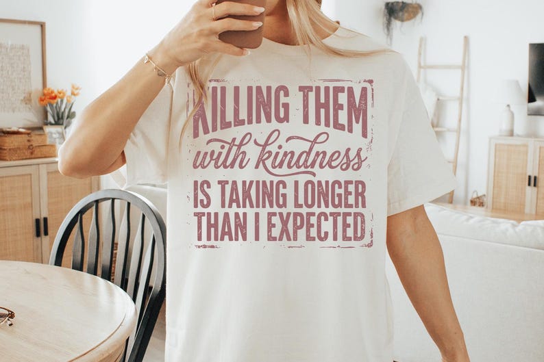 K&ouml;nnte beinhalten: Cremefarbenes T-Shirt mit dem Text "Killing them with kindness is taking longer than I expected" in einer verblassten, malvenfarbenen Schrift. Ein l&auml;ssiges Kleidungsst&uuml;ck f&uuml;r den Alltag.