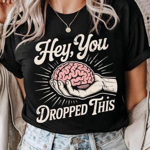 May include: Black t-shirt with the text "Hey, You Dropped This." The shirt features a graphic of a pink brain held in a hand, surrounded by radiating lines. A casual, everyday tee.