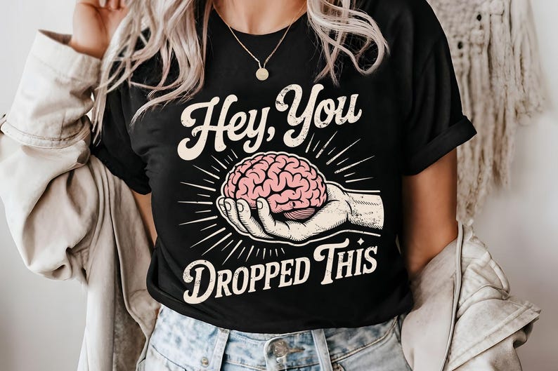 May include: Black t-shirt with the text "Hey, You Dropped This." The shirt features a graphic of a pink brain held in a hand, surrounded by radiating lines. A casual, everyday tee.