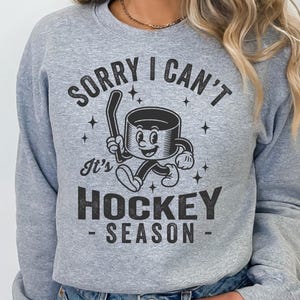May include: A pale grey sweatshirt with the text "SORRY I CAN'T It's HOCKEY SEASON" in black. The graphic shows a cartoon hockey puck character holding a hockey stick. The design includes stars.