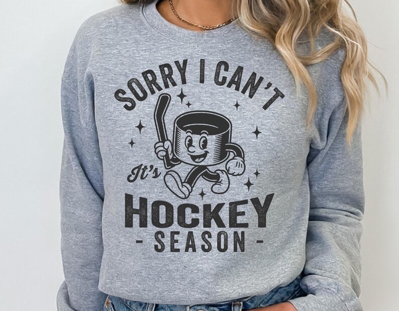 May include: A pale grey sweatshirt with the text "SORRY I CAN'T It's HOCKEY SEASON" in black. The graphic shows a cartoon hockey puck character holding a hockey stick. The design includes stars.