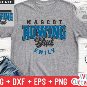 May include: A grey t-shirt with a blue and black graphic that says "MASCOT ROWING Dad EMILY". The graphic is in a varsity style font. The t-shirt is on a wooden surface with a pair of jeans and trainers.