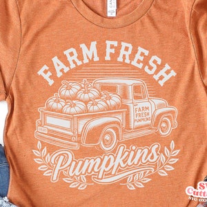 Puede incluir: Un diseño gráfico blanco sobre una camiseta naranja que representa un camión vintage lleno de calabazas. El texto dice "Farm Fresh Pumpkins".