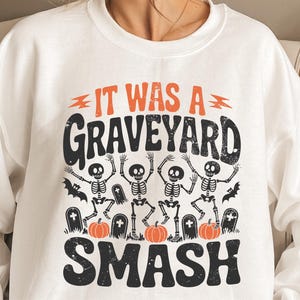May include: White sweatshirt with a Halloween-themed graphic. The design features the text "IT WAS A GRAVEYARD SMASH" in orange and black, along with dancing skeletons, pumpkins, and tombstones. A fun and festive apparel item.