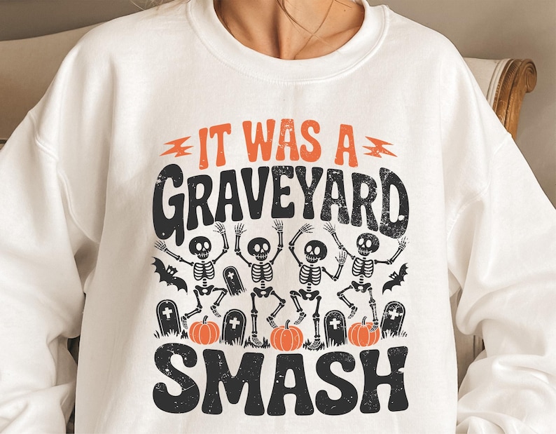 May include: White sweatshirt with a Halloween-themed graphic. The design features the text "IT WAS A GRAVEYARD SMASH" in orange and black, along with dancing skeletons, pumpkins, and tombstones. A fun and festive apparel item.