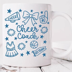 May include: White ceramic mug with a blue design featuring cheerleading-related graphics and text. The design includes a megaphone, bow, pom-poms, a jersey, a basketball, stars, and the words "Cheer Coach" and "Go Team".