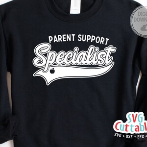 May include: Black sweatshirt with white text that reads "Parent Support Specialist" with a white apple graphic and a white baseball-style arc above the text. The text "SVG Cuttables" is in the lower right corner of the image.