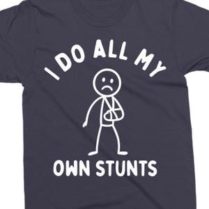 May include: Dark navy blue t-shirt with the text "I DO ALL MY OWN STUNTS" in white. The graphic features a stick figure with a sad face and a broken arm in a cast.