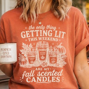 May include: A burnt orange t-shirt featuring a white graphic print of three candles and pumpkins with the text: the only thing GETTING LIT THIS WEEKEND ARE MY fall scented CANDLES. The candles are labeled Pumpkin Spice, Autumn Leaves, and Cozy Nights.