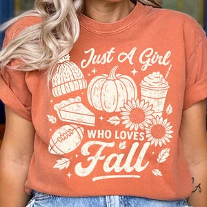 May include: Orange-colored t-shirt with the text "Just A Girl Who Loves Fall" and fall-themed graphics, including a pumpkin, sunflowers, and a football. The shirt has short sleeves and a relaxed fit.