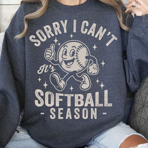 Op de afbeelding: Donkergrijze sweatshirt met de tekst "SORRY I CAN'T It's SOFTBALL SEASON" in een distressed, vintage stijl. De afbeelding toont een honkbalpersonage dat een duim omhoog geeft. De sweatshirt is een casual, comfortabel kledingstuk.
