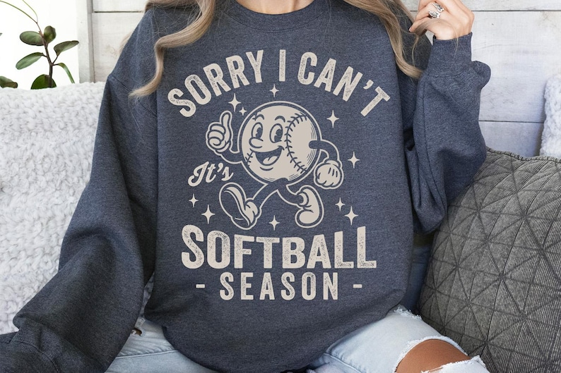 Op de afbeelding: Donkergrijze sweatshirt met de tekst "SORRY I CAN'T It's SOFTBALL SEASON" in een distressed, vintage stijl. De afbeelding toont een honkbalpersonage dat een duim omhoog geeft. De sweatshirt is een casual, comfortabel kledingstuk.