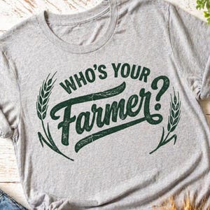 May include: A heather gray t-shirt with the text "WHO'S YOUR Farmer?" in dark green, surrounded by wheat stalks. The shirt has a crew neck and short sleeves, perfect for casual wear.