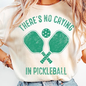 Puede incluir: Camiseta color crema con el texto "THERE'S NO CRYING IN PICKLEBALL" en verde. El diseño presenta dos palas de pickleball verdes cruzadas y una pelota de pickleball verde. Una camiseta casual y cómoda.