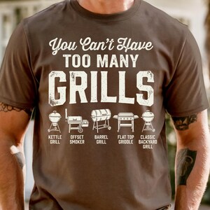 May include: A brown t-shirt with the text "You Can't Have TOO MANY GRILLS" in white. Below the text are illustrations of different grill types, including kettle, offset smoker, barrel, flat top griddle, and classic backyard grills.