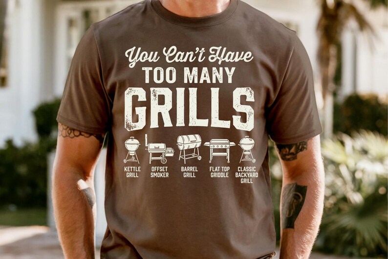 May include: A brown t-shirt with the text "You Can't Have TOO MANY GRILLS" in white. Below the text are illustrations of different grill types, including kettle, offset smoker, barrel, flat top griddle, and classic backyard grills.