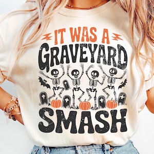 May include: Cream-colored t-shirt with a Halloween-themed graphic. The design features the text "IT WAS A GRAVEYARD SMASH" in orange and black, along with dancing skeletons, pumpkins, and tombstones. A fun and festive design.